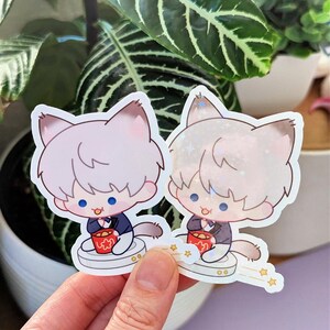 May include: Two cat-themed stickers. Each sticker features a cartoon cat character with light-colored fur, wearing a dark blue suit and holding a red cup. The stickers have a white border and are set against a background of green plants.