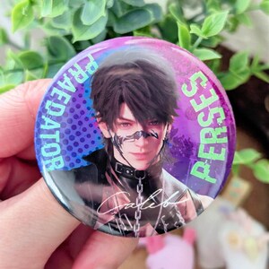 May include: A round button with a portrait of a person with dark hair and a black choker. The button has the words "PREDATOR" and "PERSES" in a purple font around the edge. The person's signature is visible.