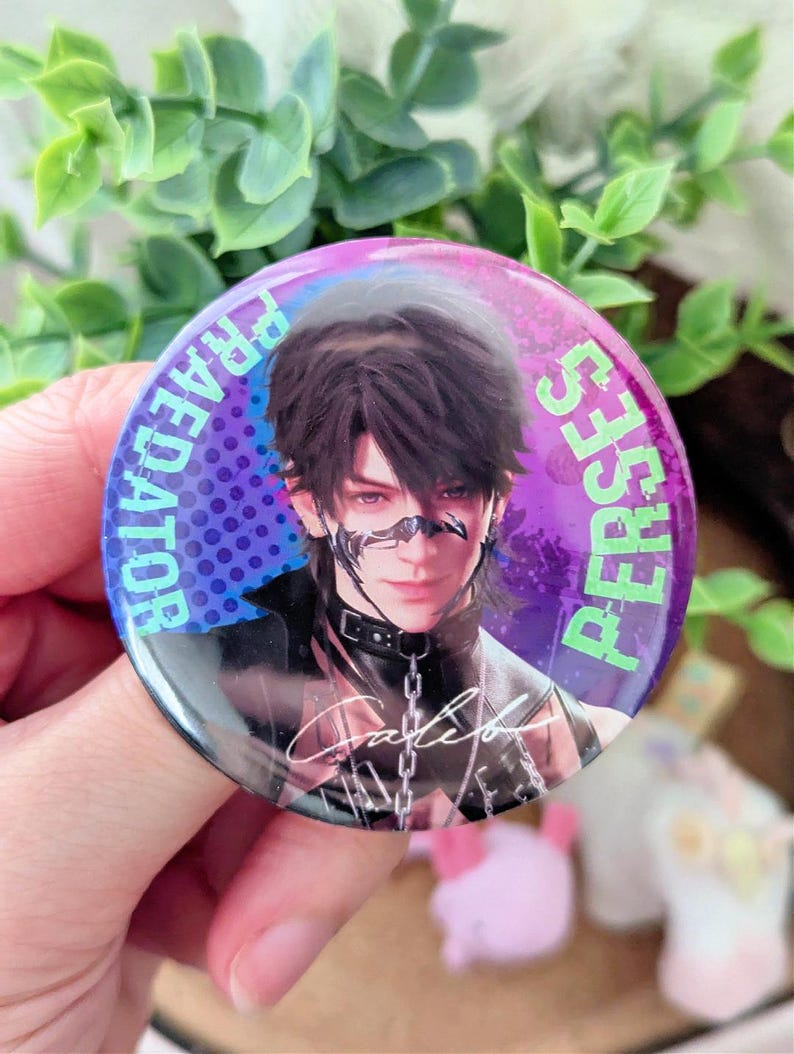 May include: A round button with a portrait of a person with dark hair and a black choker. The button has the words "PREDATOR" and "PERSES" in a purple font around the edge. The person's signature is visible.
