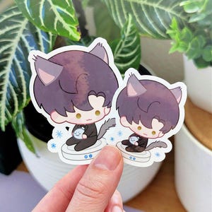 May include: Two stickers featuring a cartoon character with dark purple hair, cat ears, and a gray tail. The character is wearing a black suit and holding a small, round object. The stickers are on a white background with blue snowflake accents.