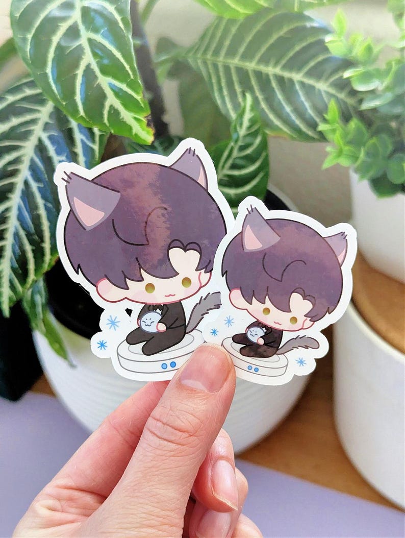 May include: Two stickers featuring a cartoon character with dark purple hair, cat ears, and a gray tail. The character is wearing a black suit and holding a small, round object. The stickers are on a white background with blue snowflake accents.