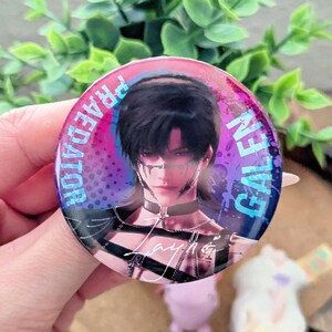 May include: A round button with a portrait of a person with black hair and a choker. The button has the words "GALEN" and "PREDATOR" in blue and white text around the edge. The background is a mix of pink, purple, and blue.
