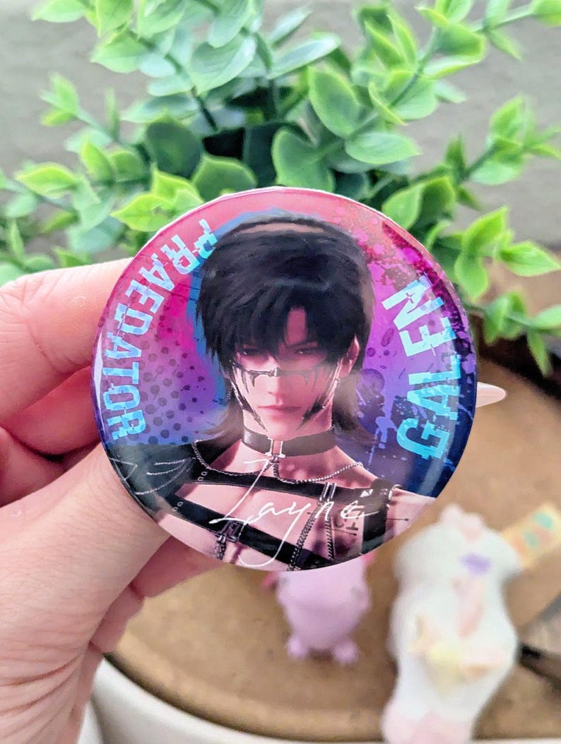 May include: A round button with a portrait of a person with black hair and a choker. The button has the words "GALEN" and "PREDATOR" in blue and white text around the edge. The background is a mix of pink, purple, and blue.