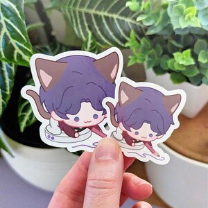 May include: Two stickers featuring a cartoon character with purple hair and cat ears. The character is wearing a maroon outfit and is seated in a white cup. The stickers are held in a hand in front of a green leafy background.