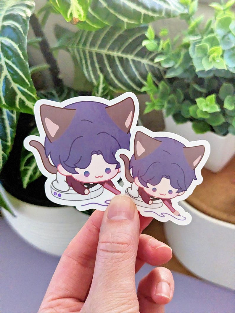 May include: Two stickers featuring a cartoon character with purple hair and cat ears. The character is wearing a maroon outfit and is seated in a white cup. The stickers are held in a hand in front of a green leafy background.