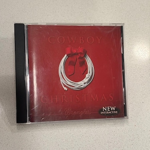 Puede incluir: Una caja de CD con una cubierta roja con las palabras "COWBOY CHRISTMAS" y un diseño de lazo de cuerda blanca con un lazo rojo. La caja del CD está en una caja de plástico transparente.