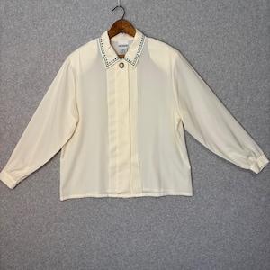 Puede incluir: Blusa crema de manga larga con cuello. El cuello está adornado con pequeños puntos verdes. La blusa tiene cierre de botones y paneles verticales en la parte delantera. La prenda está colgada en una percha de madera.