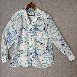 Könnte beinhalten: Weißes Button-Down-Hemd mit floralem Muster in Blau-, Türkis- und Schwarztönen. Das langärmlige Hemd hängt an einem Holzbügel. Das florale Design umfasst stilisierte Pflanzen- und Blattmotive.