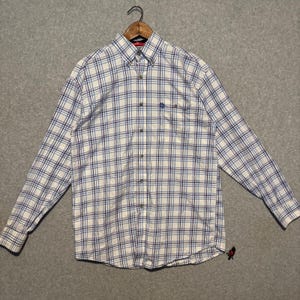 May include: A long-sleeved shirt with a white and blue check pattern, hanging on a wooden hanger. The shirt has a button-down collar and a chest pocket. The background is plain grey. The shirt has a small logo on the pocket.