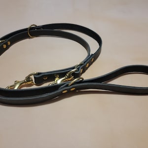 May include: A black leather dog lead with two brass clasps and a brass ring. The lead is doubled over and has a loop at the end.