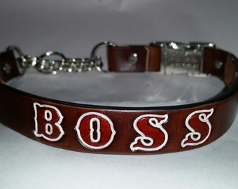 Può includere: Un collare per cani in pelle marrone con una fibbia argentata e una catena. Il collare ha la parola "BOSS" impressa in lettere rosse e bianche.