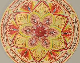 Puede incluir: Mandala redondo pintado a mano con un diseño floral. La obra de arte presenta una flor roja central rodeada de formas de pétalos amarillos, naranjas y rojos. El borde exterior tiene un borde decorativo con pequeños puntos y patrones en forma de hojas. Adornado con pequeños acentos brillantes.