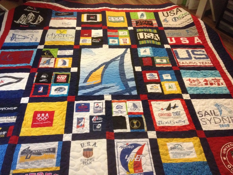 May include: A colourful quilt made of fabric squares with various sailing-themed logos and designs. The quilt features a large central square with a sailboat design and the words "USA" and "Sailing Team".