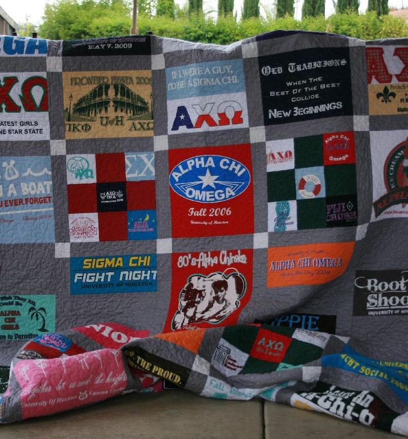 May include: A patchwork quilt made from t-shirts with various college logos and Greek letters. The quilt is made in a grid pattern with a grey background. The quilt is folded in half and lying on a brown surface.