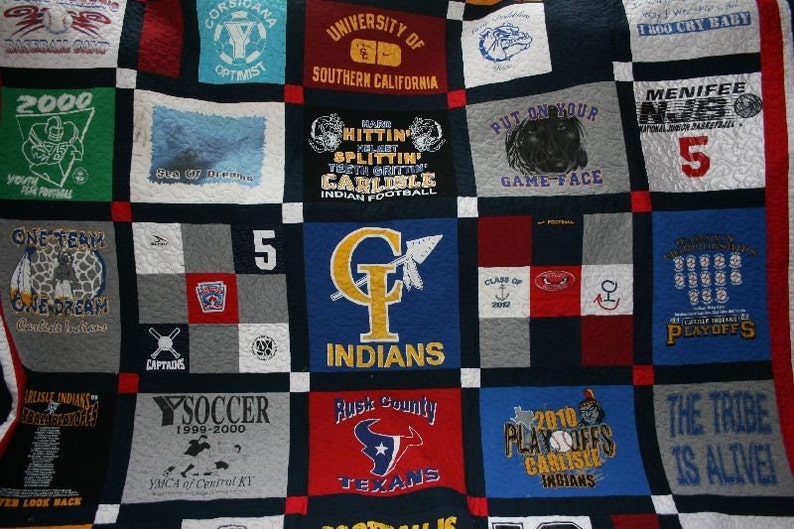 May include: A quilt made from a variety of t-shirts, featuring different logos and designs. The quilt is made up of squares, each with a different design. Some of the designs include the words "One Team One Dream", "Put On Your Game Face", and "The Tribe Is Alive!"