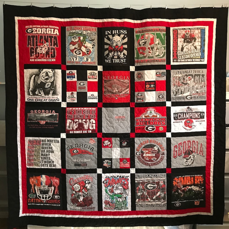 May include: A quilt made from University of Georgia t-shirts. The quilt is black, red, white, and gray. It features the school's mascot, the bulldog, and the team's logo. The quilt is made up of 25 squares, each with a different design. The quilt is a great way to show your school spirit.