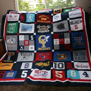 May include: A blue and red patchwork quilt made from t-shirts with various sports team logos and numbers. The quilt is made up of 25 squares, each with a different design. Some of the designs include the words "COSMOS", "INDIANS", "THE TRIBE IS ALIVE", and "BELIEVING ACHIEVING".