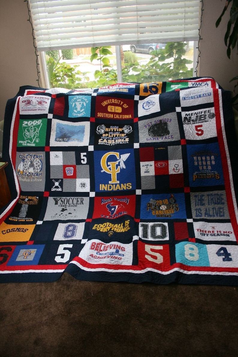 May include: A blue and red patchwork quilt made from t-shirts with various sports team logos and numbers. The quilt is made up of 25 squares, each with a different design. Some of the designs include the words "COSMOS", "INDIANS", "THE TRIBE IS ALIVE", and "BELIEVING ACHIEVING".