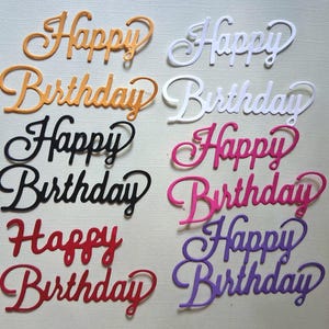 May include: A collection of colorful, cursive "Happy Birthday" cutouts. The words are in various colors, including orange, white, black, pink, red, and purple. These decorative elements are ideal for crafting and card making.