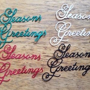 May include: Four die-cut paper embellishments with the words "Seasons Greetings" in a cursive font. The embellishments are in green, white, red, and black.