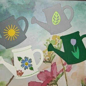 May include: Four decorative watering cans in various colors. One gray can features a yellow sun, another gray can has a green leaf, a white can has a blue flower, and a green can has a purple tulip. The cans are likely paper cutouts.