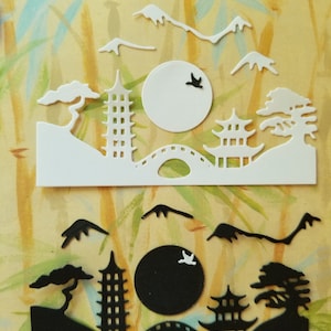 May include: Two die-cut paper designs of a Japanese landscape with a pagoda, bridge, mountains, trees, and a moon. The top design is white and the bottom design is black.