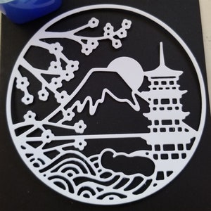 May include: White paper die cut of a Japanese scene with Mount Fuji, a pagoda, cherry blossoms, and waves. The die cut is circular and is on a black background.