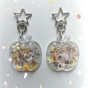 Może przedstawiać: Dwa przezroczyste breloczki do kluczy w kształcie jabłka z zapięciami w kształcie gwiazdy. Każdy breloczek zawiera kolorowy obrazek postaci w stylu anime, wraz z małymi elementami dekoracyjnymi. Widoczne jest słowo "cutie".