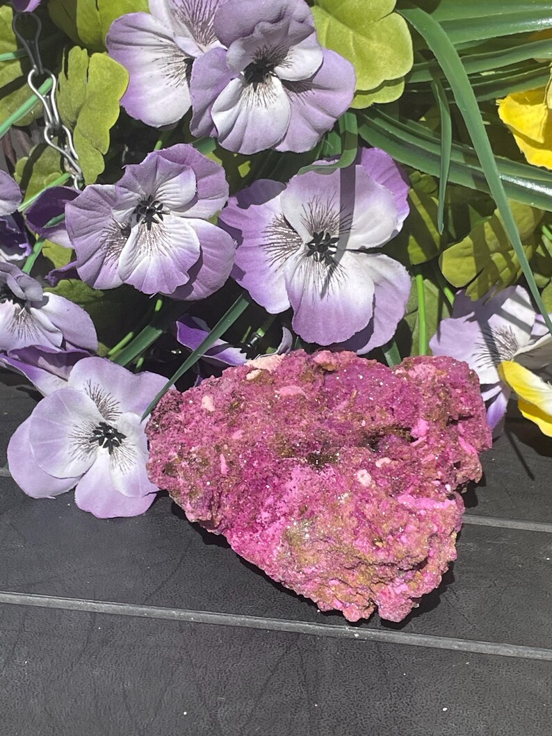 May include: A striking pink mineral with a rugged, textured appearance. Artificial purple and white pansy flowers with green leaves surround the mineral, providing a vivid contrast. The background is a dark surface.