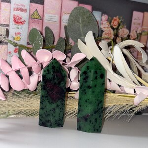 May include: Two green and red ruby zoisite crystal towers. The crystals have a dark green base colour with black speckles and red inclusions. The crystals are displayed with pink and white floral accents.
