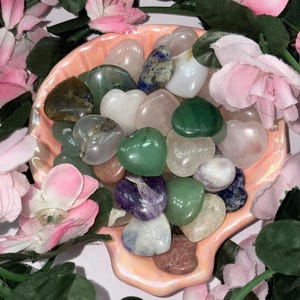 May include: A selection of heart-shaped gemstones in shades of green, pink, white, and purple, presented in a light pink, shell-shaped dish. The dish is encircled by pink and white artificial flowers and green foliage.