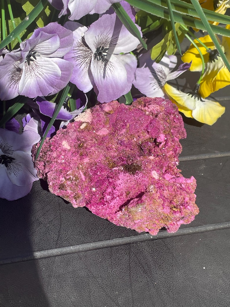 May include: A striking pink mineral specimen with a textured surface and touches of gold and brown. Artificial purple, white, and yellow flowers surround the mineral. The background is a dark, textured surface.