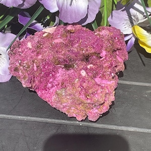 Op de afbeelding: Een levendig, getextureerd, roze mineraal specimen met hints van bruin en goud. De ruwe steen staat tegen een achtergrond van paarse en gele kunstbloemen, wat een opvallend contrast creëert. Het oppervlak van het mineraal heeft een kristallijn uiterlijk.
