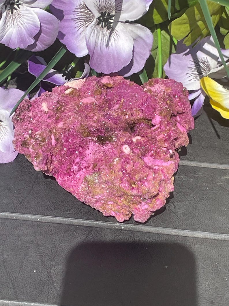 May include: A textured, pink mineral specimen with brown and gold hues. The rough stone is set against purple and yellow artificial flowers. The mineral's surface has a crystalline appearance. The image shows a close-up of the mineral.