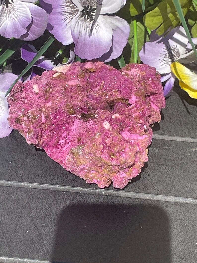 May include: A textured mineral specimen in shades of magenta, brown, and white. The rock has an irregular shape and is likely a natural formation. The background features purple and yellow flowers.