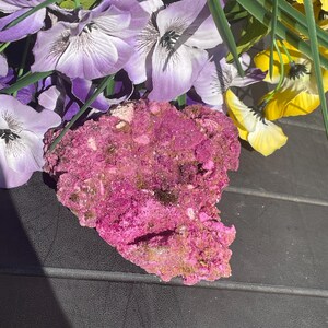 May include: A bright pink mineral specimen with a textured surface. The mineral is set against a backdrop of purple and yellow artificial flowers, creating a colourful contrast. The specimen's rough edges and varied hues suggest a natural, unrefined form.