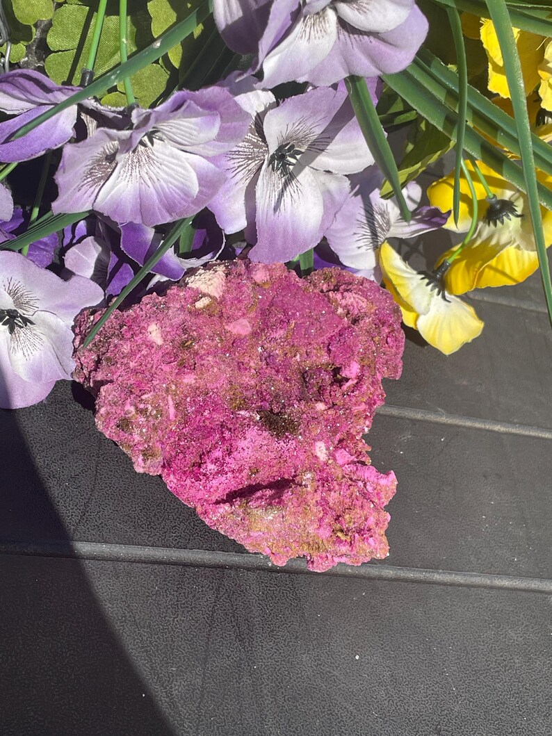 May include: A bright pink mineral specimen with a textured surface. The mineral is set against a backdrop of purple and yellow artificial flowers, creating a colourful contrast. The specimen's rough edges and varied hues suggest a natural, unrefined form.