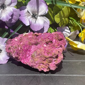 May include: A striking mineral specimen with a rough texture, predominantly a deep magenta hue with touches of gold and brown. Artificial purple, yellow, and green flowers surround the mineral, providing a vivid contrast.