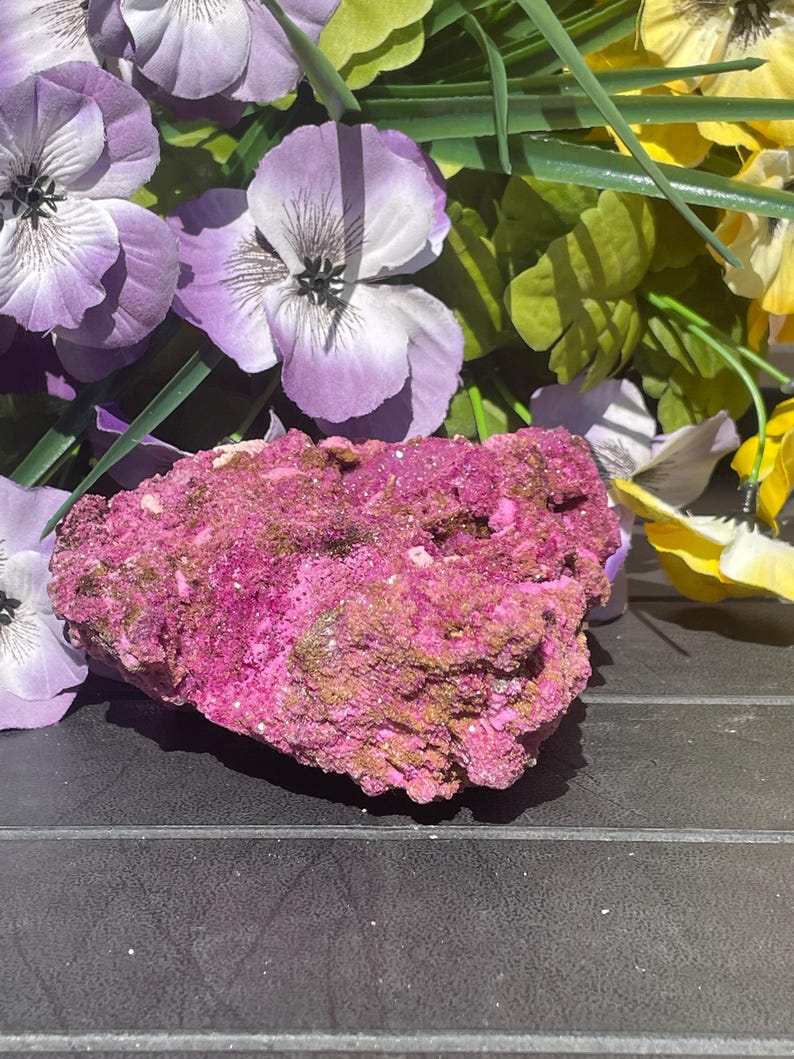 May include: A striking mineral specimen with a rough texture, predominantly a deep magenta hue with touches of gold and brown. Artificial purple, yellow, and green flowers surround the mineral, providing a vivid contrast.
