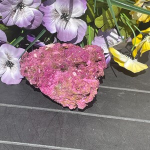 May include: A bright pink mineral specimen with a rugged, textured surface. Artificial purple and yellow flowers surround the mineral, creating a colourful contrast. The specimen is displayed on a black surface.