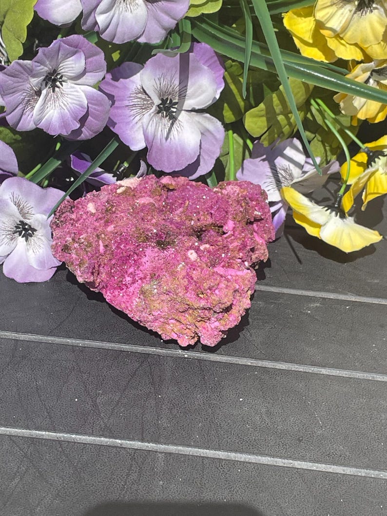 May include: A bright pink mineral specimen with a rugged, textured surface. Artificial purple and yellow flowers surround the mineral, creating a colourful contrast. The specimen is displayed on a black surface.