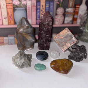 May include: A collection of various polished gemstones and crystals. The assortment includes a tall, dark purple crystal, a gray, freeform stone, and several smaller, rounded stones in shades of green, brown, and gray. A pink and white shelf is in the background.