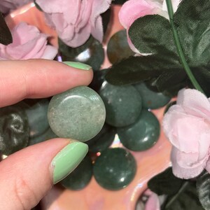 May include: A close-up of several small, round, green stones. One stone is held between fingers with light green nail polish. Pink and white flowers and green leaves are in the background. The stones have a smooth, polished surface.