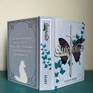 May include: A hardback book titled "Quicksilver" with a pale blue cover. The cover features a butterfly design with a sword, and the author's name, Callie Hart. The spine reads "Quicksilver" and "HART". The back cover has a quote and a white fox illustration.