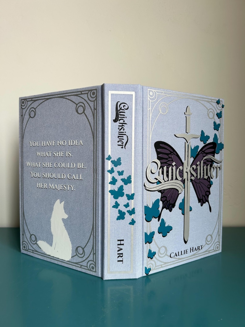 May include: A hardback book titled "Quicksilver" with a pale blue cover. The cover features a butterfly design with a sword, and the author's name, Callie Hart. The spine reads "Quicksilver" and "HART". The back cover has a quote and a white fox illustration.