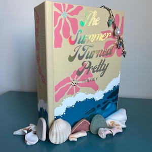 Puede incluir: Un libro titulado "The Summer I Turned Pretty Trilogy" con una cubierta color crema con diseños florales rosas y letras holográficas. Una cadena decorativa con dijes cuelga de la parte superior. Conchas marinas y piedras están dispuestas en la base.