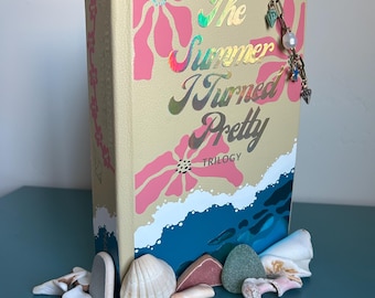 Può includere: Un libro intitolato "The Summer I Turned Pretty Trilogy" con una copertina color crema con disegni floreali rosa e scritte olografiche. Una catena decorativa con ciondoli pende dalla parte superiore. Conchiglie e pietre sono disposte alla base.