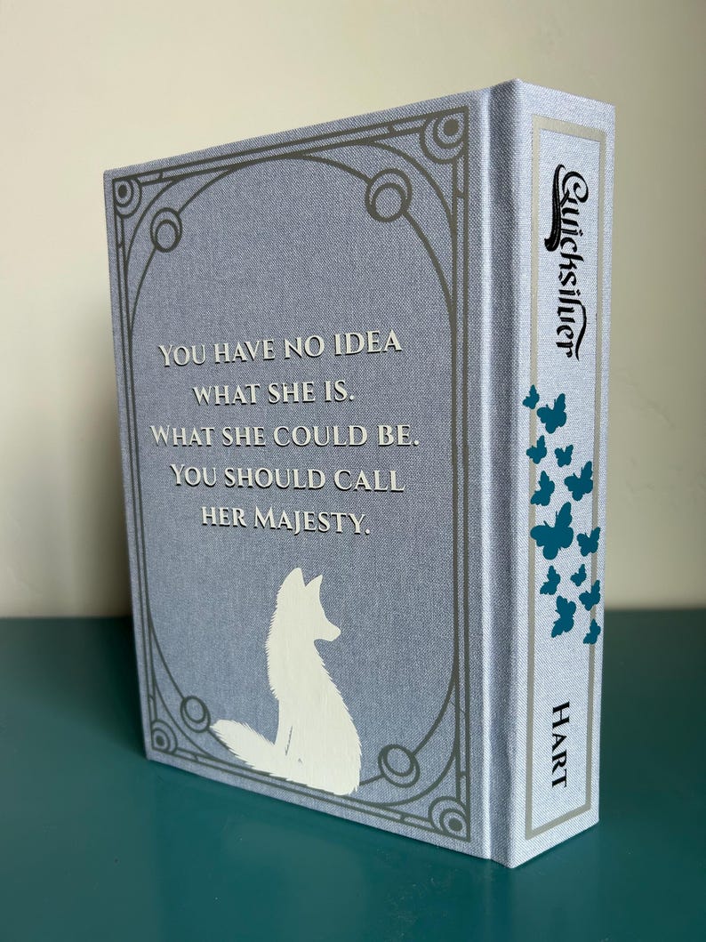 May include: A hardback book titled "Quicksilver" with a pale blue cover. The cover features a white fox silhouette and text that reads "YOU HAVE NO IDEA WHAT SHE IS. WHAT SHE COULD BE. YOU SHOULD CALL HER MAJESTY." The spine has teal butterflies.