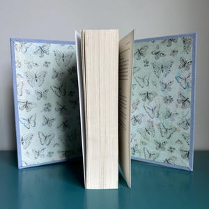 May include: A hardback book with a light blue cover adorned with a butterfly design. The book is open, displaying the pages. The cover has a light blue border. The background is a solid light blue.