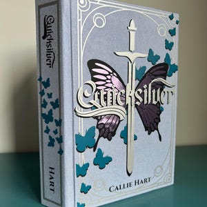 May include: A hardback book titled "Quicksilver" by Callie Hart. The cover is light blue with a silver sword and butterfly design. Teal butterflies and the author's name are also visible on the cover.